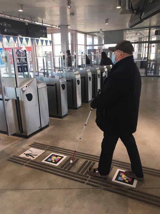User with white cane crossing the ticket gates at Gare de Versailles-Chantiers with NaviLens codes on the floor
