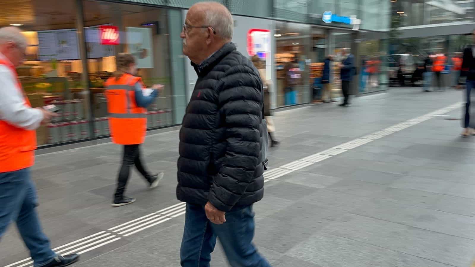 Passeggero anziano con giacca imbottita che attraversa la galleria commerciale di Den Haag Centraal sulla linea guida tattile, con una volontaria con gilet arancione di ProRail che accompagna il test in fondo