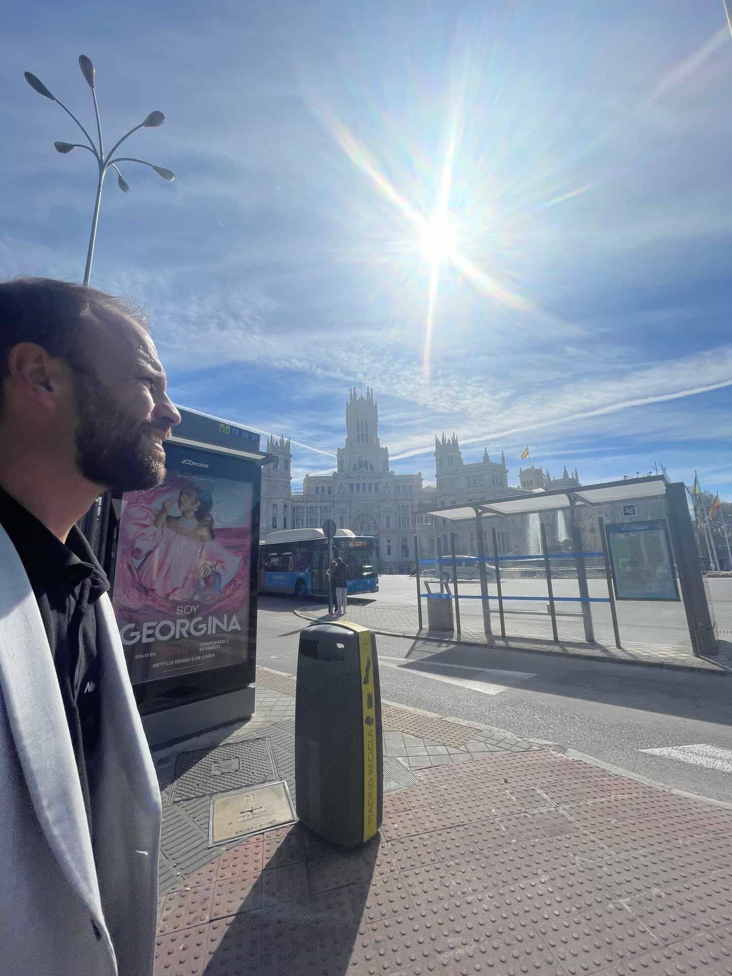 Un utente a una fermata della EMT in Plaza de Cibeles guarda verso la pensilina successiva; il sole è alto, in primo piano un MUPI pubblicitario della serie «Soy Georgina» e, in fondo, il Palacio de Comunicaciones e la fontana di Cibele