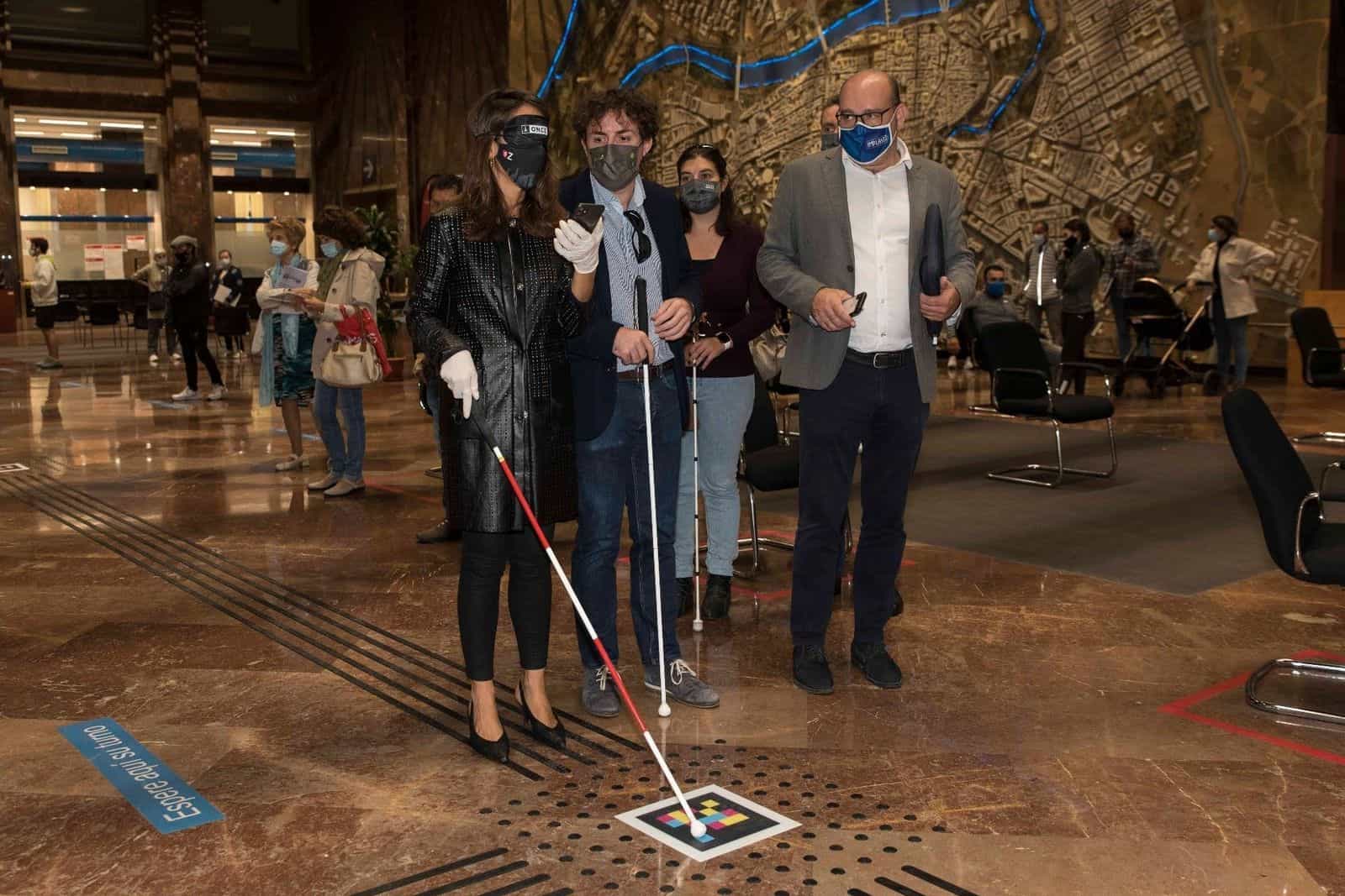 Un utente della ONCE con bastone bianco mostra l’app NaviLens sul proprio telefono all’assessora María Navarro, che ascolta con la mascherina oscurante mentre tiene il proprio bastone; in fondo alla hall del Comune, un’altra utente con mascherina e bastone partecipa alla dimostrazione