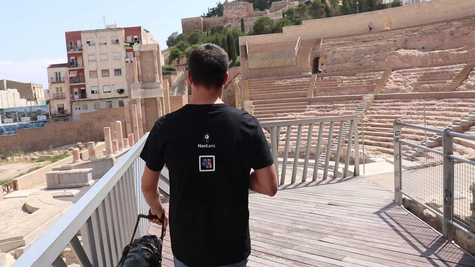 Nutzer mit NaviLens-T-Shirt auf dem Außensteg des MUSTER mit Panoramablick auf das Römische Theater von Cartagena im Hintergrund