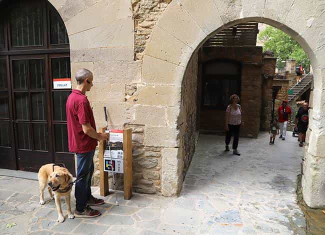 A visitor with a yellow Labrador guide dog and a white cane, standing at an interpretive panel on wooden posts carrying its NaviLens code, under the fortified gateway of Sant Miquel del Fai; behind them, the cobbled courtyard and other visitors