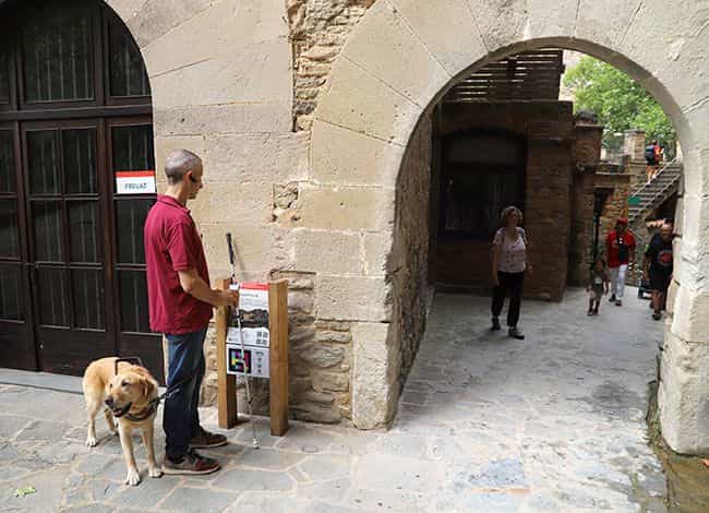 Ein Besucher mit gelbem Labrador-Blindenführhund und weißem Langstock vor einer Informationstafel auf Holzpfosten mit ihrem NaviLens-Code, unter dem befestigten Tor von Sant Miquel del Fai; dahinter der gepflasterte Hof und weitere Besucherinnen und Besucher