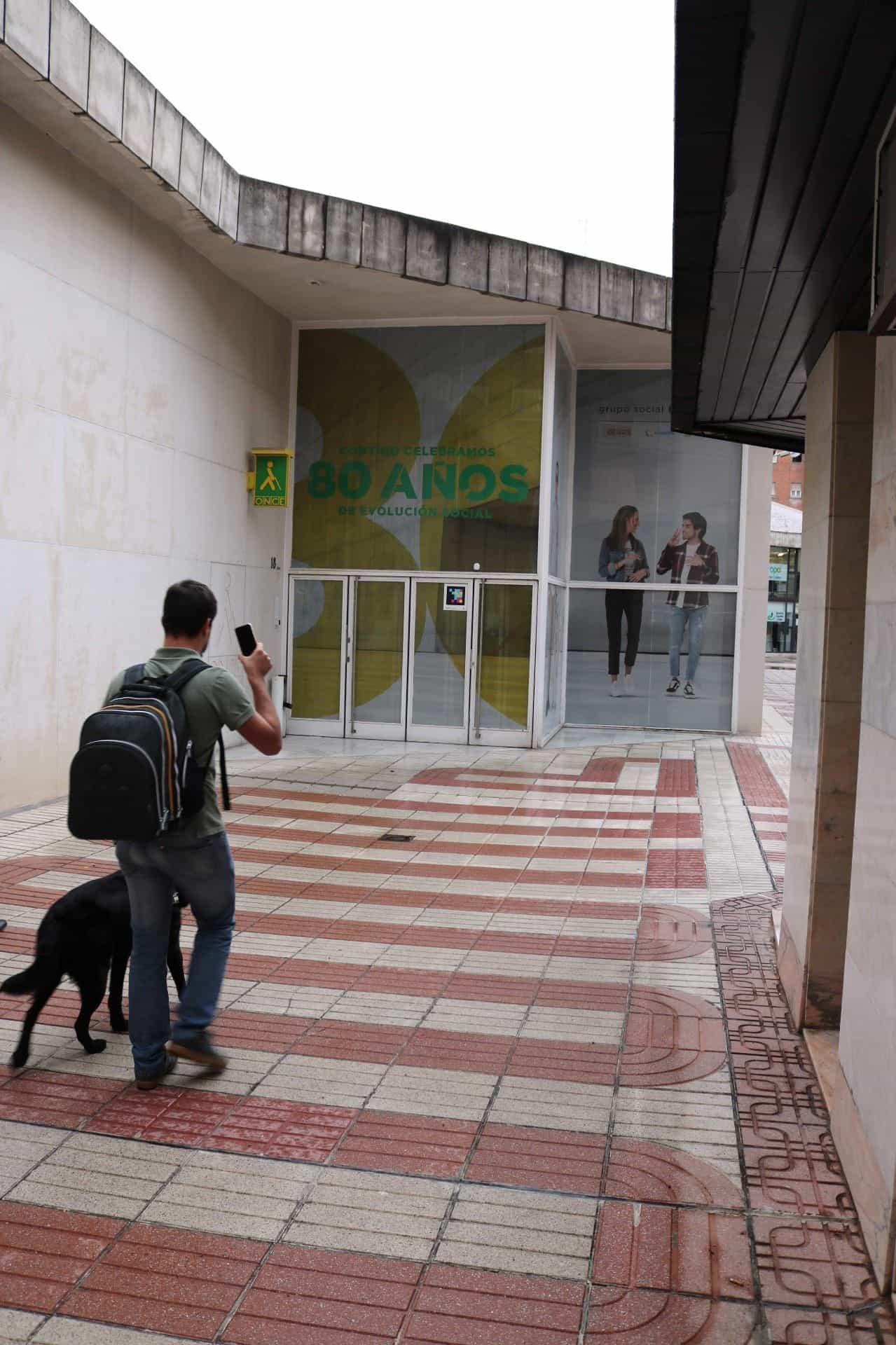 Giovane con zaino e cane guida nero che inquadra con il telefono il codice NaviLens collocato accanto alla porta a vetri d’ingresso della Delegazione dell’ONCE a Valladolid; in fondo, il telone commemorativo «80 años de evolución social» e l’insegna luminosa giallo e verde del logo ONCE su un pavimento con fasce tattili rosse e bianche