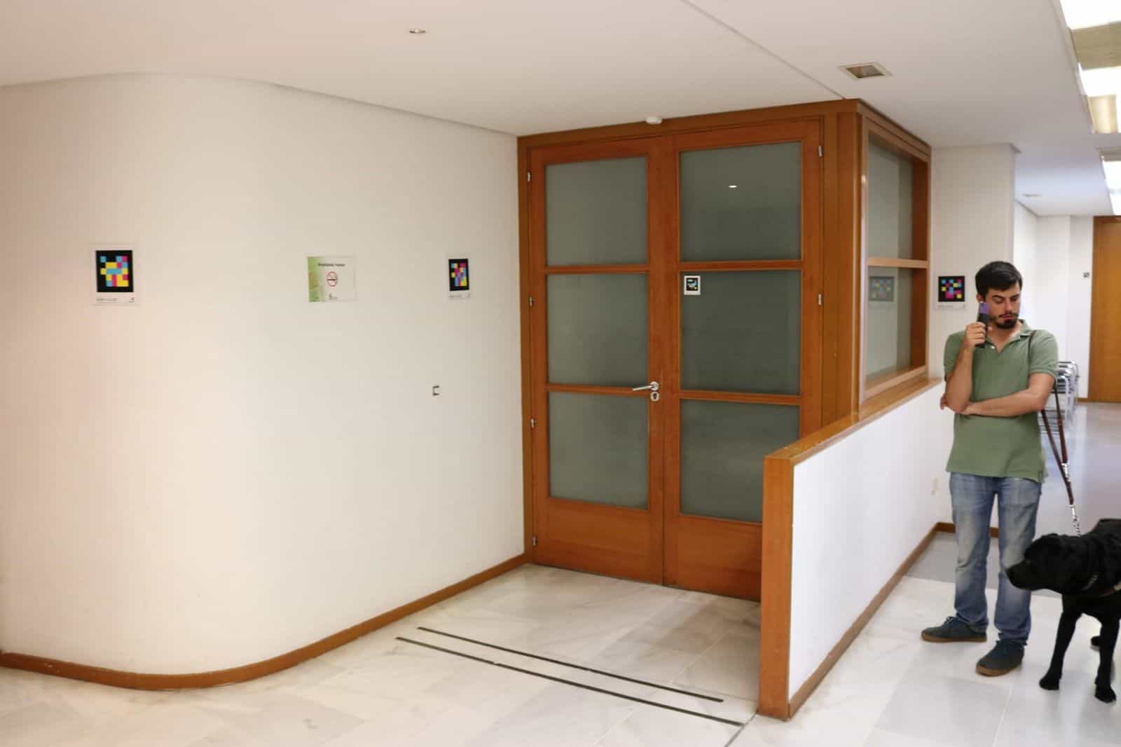 Corridoio bianco dell’ONCE Valladolid con vari codici NaviLens incorniciati sulla parete, una porta di legno con vetri satinati e, in primo piano a destra, un giovane utente con cane guida nero che consulta il telefono dopo aver inquadrato i codici