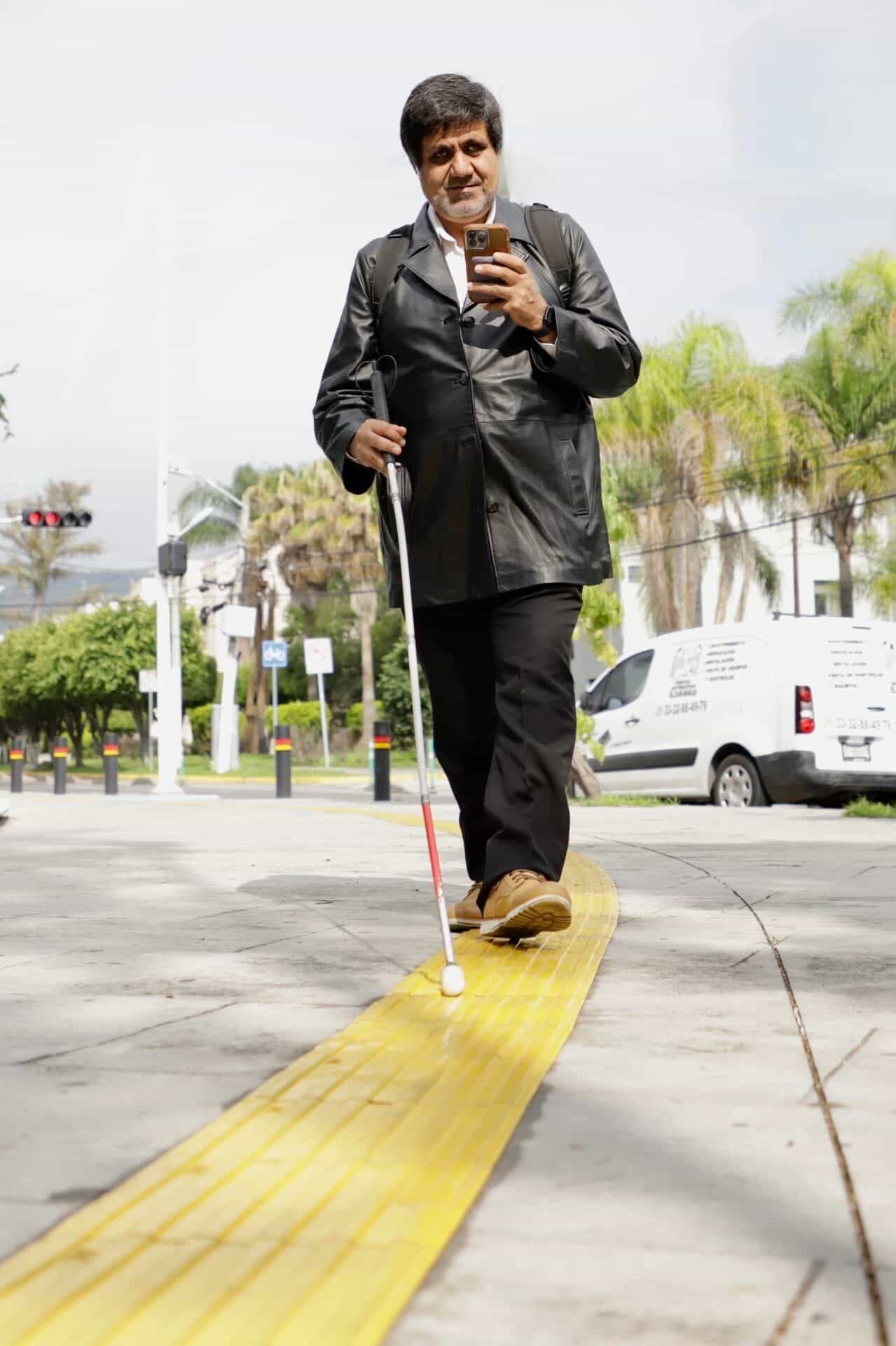 Man with a red-and-white cane walking along a long yellow tactile strip while checking the NaviLens app on his iPhone in front of the Zapopan Foreign Affairs building