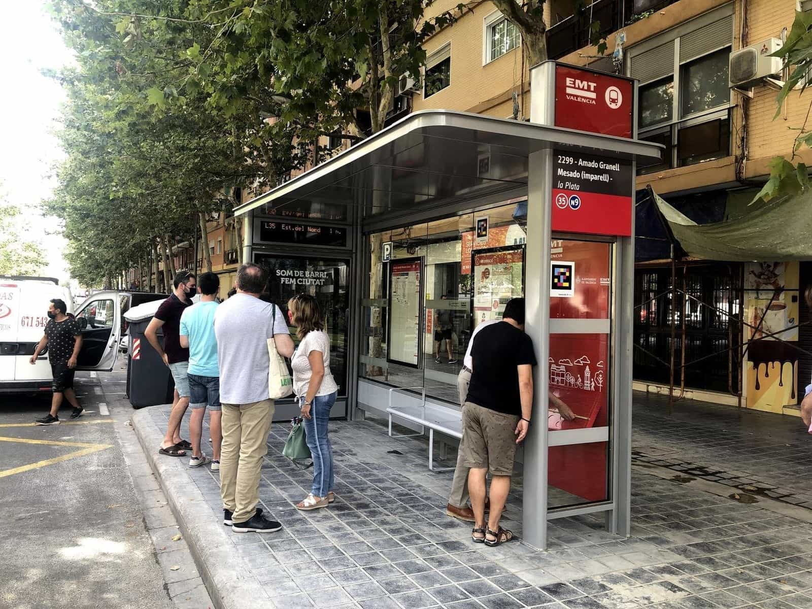 Nuova pensilina della EMT València alla fermata 2299 «Amado Granell Mesado» nel quartiere di Monteolivet, su un viale alberato; alcune persone aspettano il bus accanto al pannello «Som de barri, fem ciutat» e, sul fianco rosso del modulo, si vede un codice NaviLens quadrato a colori