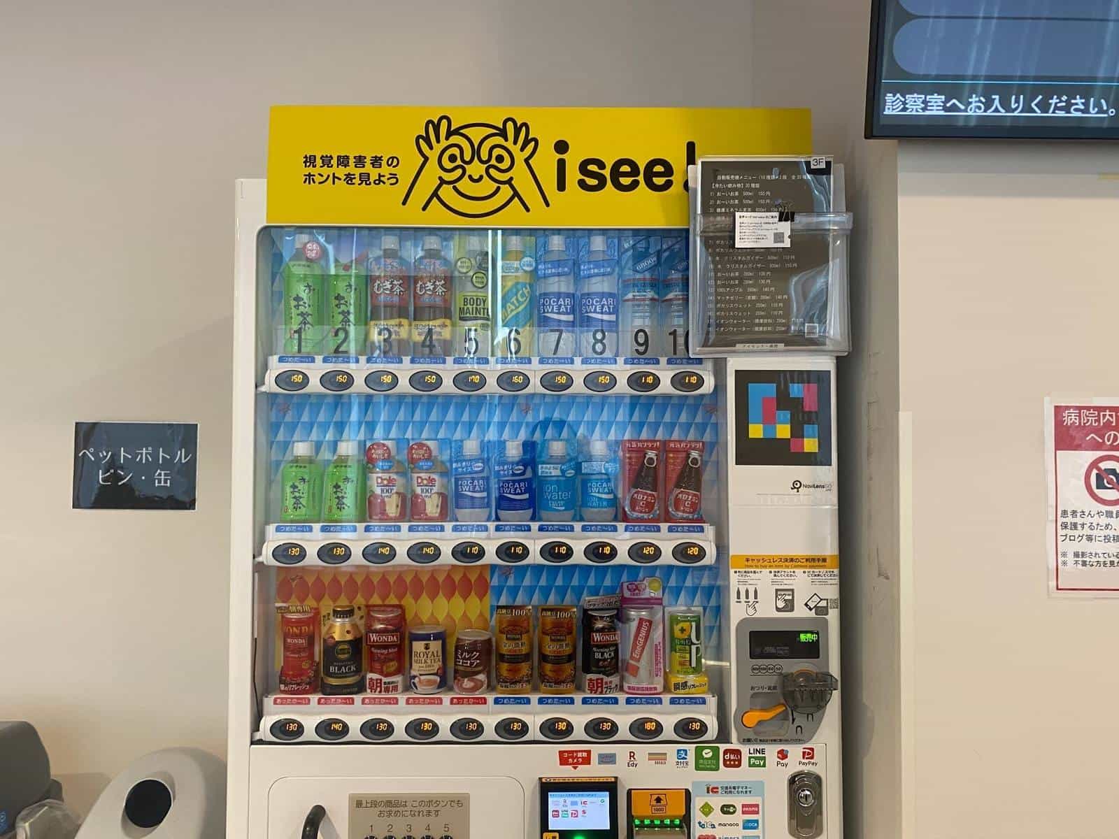 isee! vending machine with a yellow “視覚障害者のホントを見よう / i see!” poster and a large NaviLens GO tag on the right side — a blind person can hear brand, flavour, price and position of each drink right from their cane