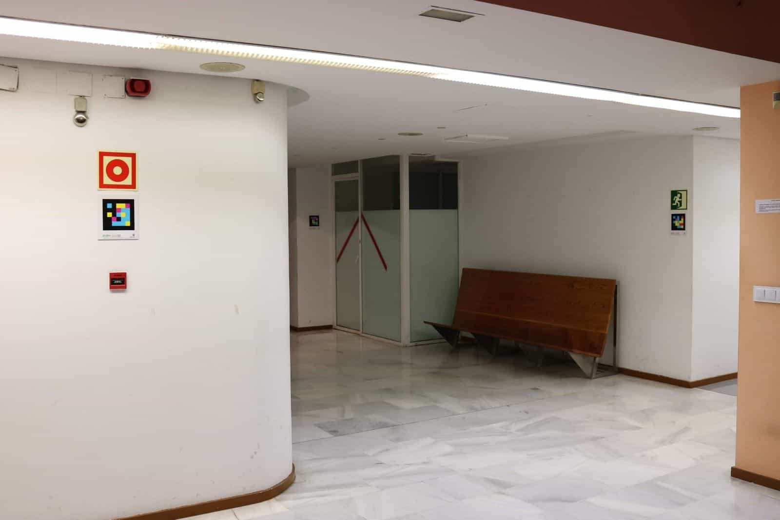 Atrio interno dell’ONCE Valladolid con pavimento di marmo bianco: su una colonna circolare, un idrante rosso con un codice NaviLens quadrato a colori sotto; in fondo, un altro codice accanto a una porta a vetri con nastri adesivi rossi e un cartello verde di uscita di emergenza sulla parete laterale destra