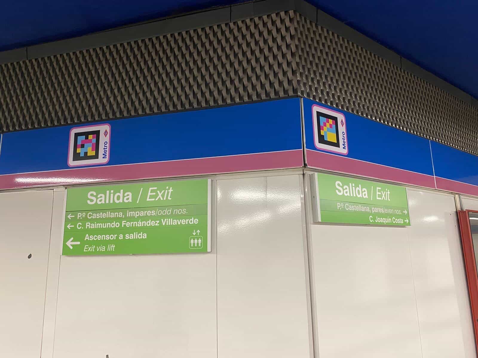 Verteilerebene der Station Nuevos Ministerios der Metro de Madrid: zwei blinde Nutzer mit Blindenstock posieren neben einem NaviLens-Techniker auf einem Blindenleitstreifen; im Boden markiert ein quadratischer, farbiger NaviLens-Code den Punkt. Im Hintergrund rot-blaue Metro-Fahrkartenautomaten und Ausgangsschilder Richtung Nuevos Ministerios und Vorortbahn