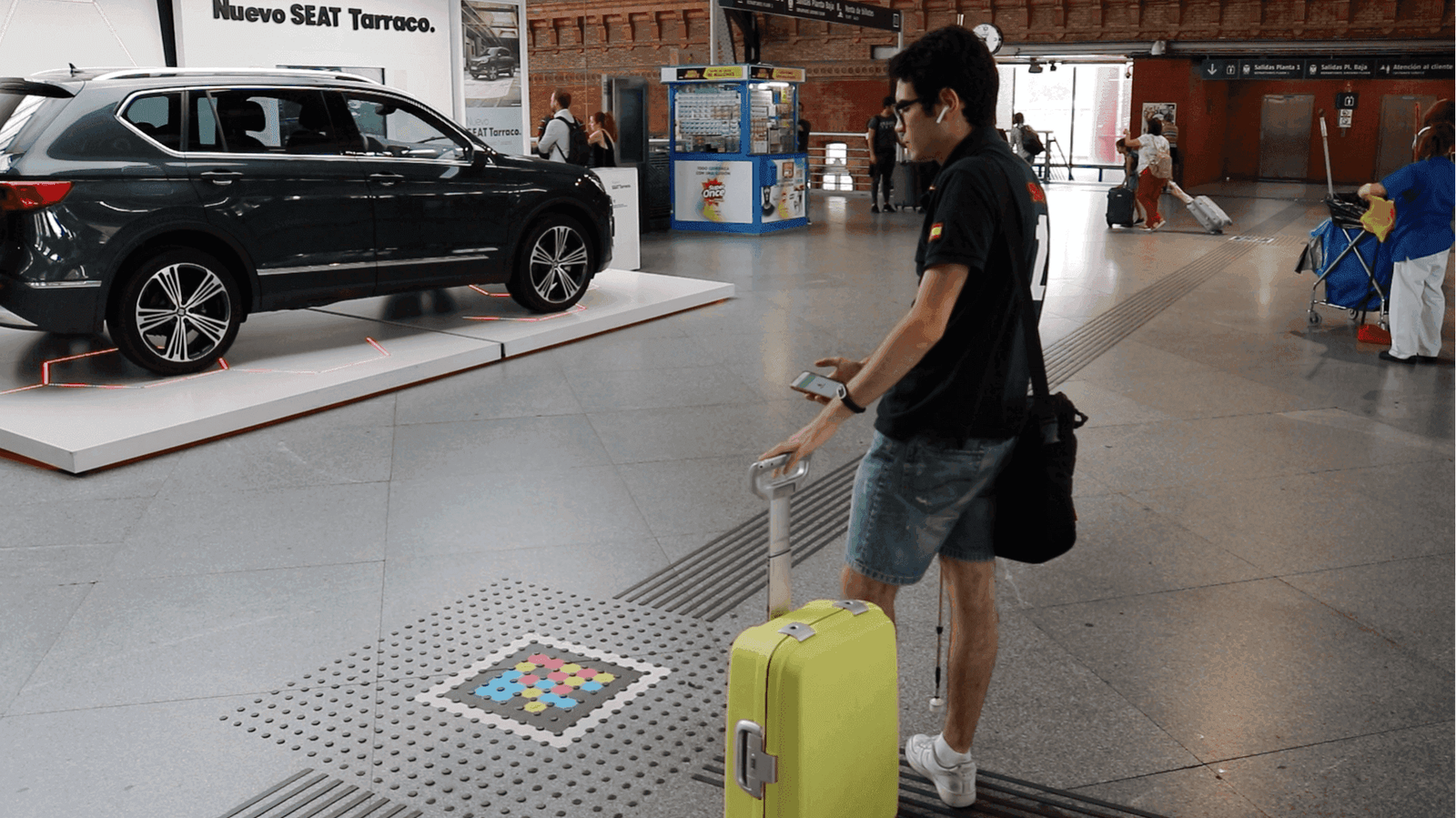 Atocha concourse next to the SEAT Tarraco display: a traveler with a yellow suitcase and headphones approaches a colorful square NaviLens code set into a tactile paving strip on the floor, checking the app on their phone.