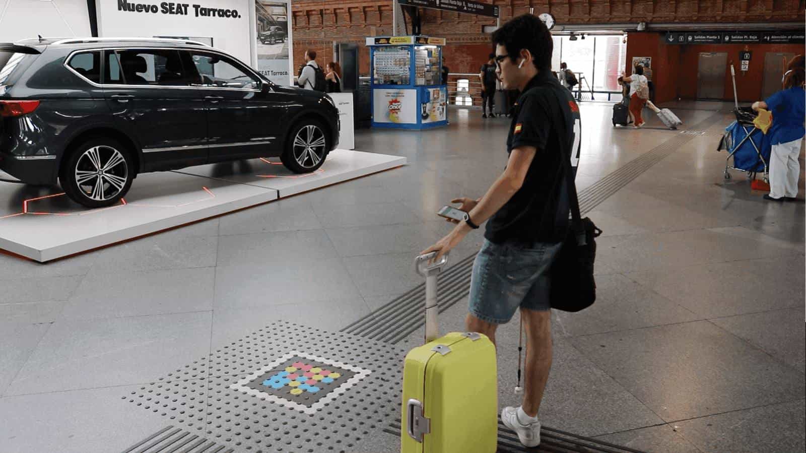 Atocha concourse next to the SEAT Tarraco display: a traveller with a yellow suitcase and headphones approaches a colourful square NaviLens code set into a tactile paving strip on the floor, checking the app on their phone.