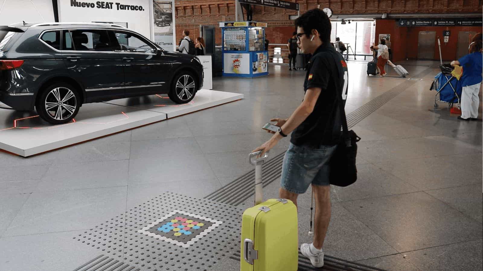 Atrio di Atocha accanto all’espositore della SEAT Tarraco: un viaggiatore con valigia gialla e cuffie si avvicina a un codice NaviLens quadrato a colori integrato in una fascia tattile del pavimento, mentre consulta l’app sul telefono