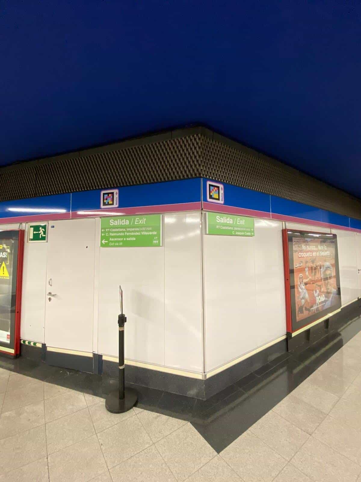 Wide view of the same Nuevos Ministerios concourse corner, with the NaviLens codes high on the blue edge, the green Exit signs towards Castellana and Joaquín Costa, a service door and an advertising sign in the side corridor