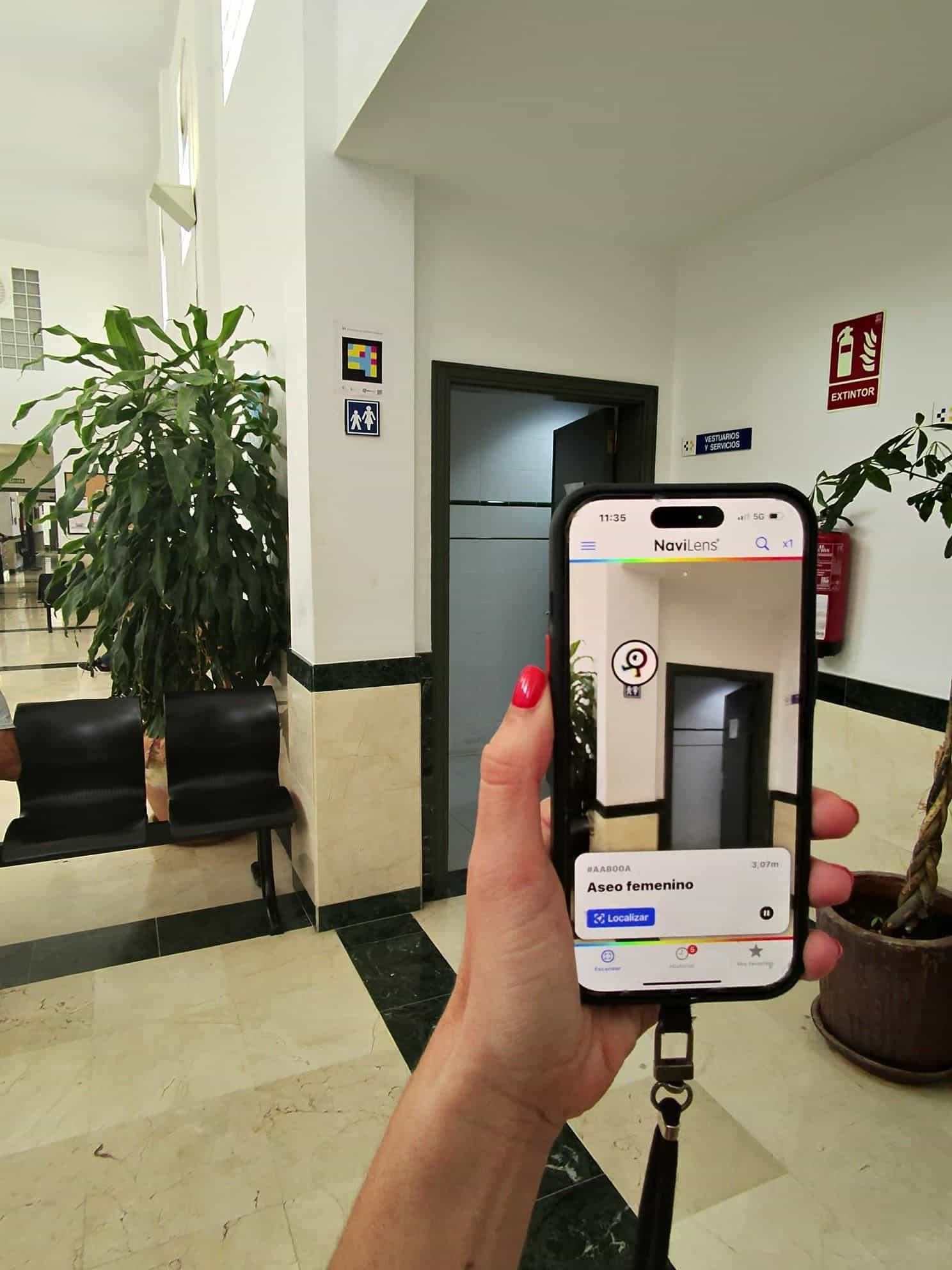 観葉植物、待合ベンチ、トイレのピクトグラム、壁のNaviLensコードがある保健センターの歴史的ホール。アプリは「#AAB00A・女性トイレ・3.07 m」を表示している。