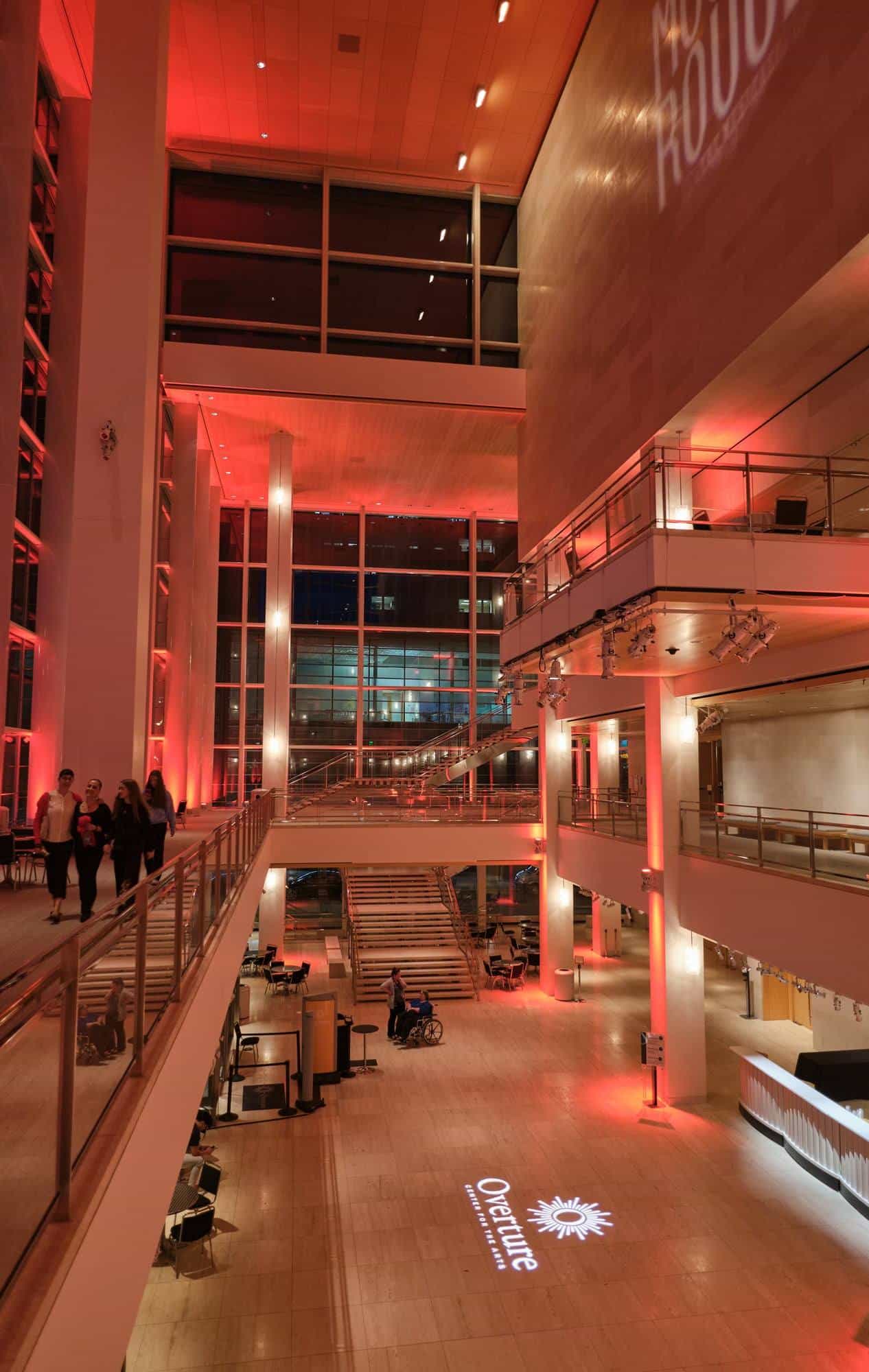 View of the Overture Center atrium from the upper gallery: a guest in a wheelchair talks with another person next to the staircase, with the Overture logo projected on the stone floor
