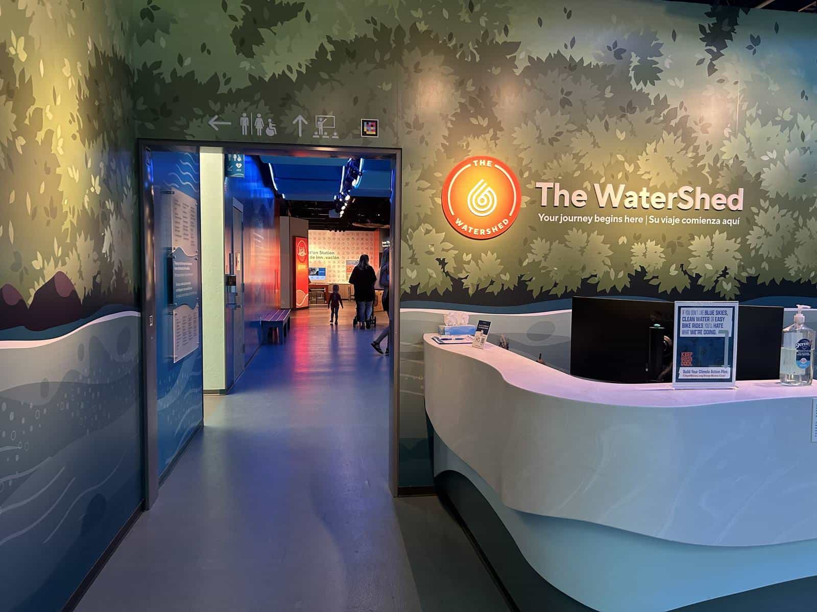 The WaterShed lobby: the centre's lit sign over the leaf mural, with the bilingual line “The WaterShed — Your journey begins here / Su viaje comienza aquí”, the front desk on the right and, through the doorway, visitors walking into the exhibit hall