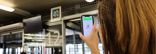 A traveler points her phone at a NaviLens code placed above the platform 12 sign at Atocha; the phone screen shows the NaviLens logo and a green message with the boarding information.