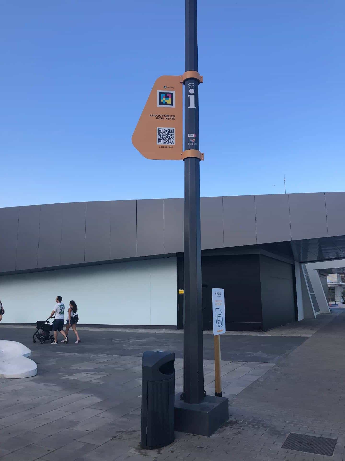 Outside esplanade of Vialia Vigo intermodal station with a black smart-city post carrying an orange plate with the NaviLens GO logo, its colour code and the access QR to the Smart Public Space; in the background, a family walks with a pushchair next to a 'Bus station' sign