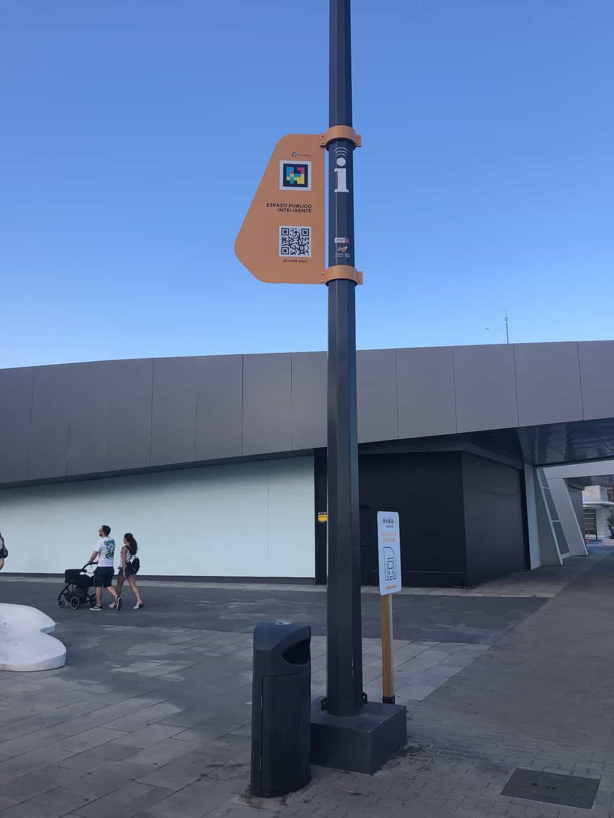 Explanada exterior de la estación intermodal Vialia Estación de Vigo con un poste negro de smart city sobre el que cuelga una placa naranja con el logo NaviLens GO, su código a colores y el QR de acceso al Espazo Público Intelixente; al fondo, una familia camina con cochecito y un cartel señaliza la 'Estación de autobuses'