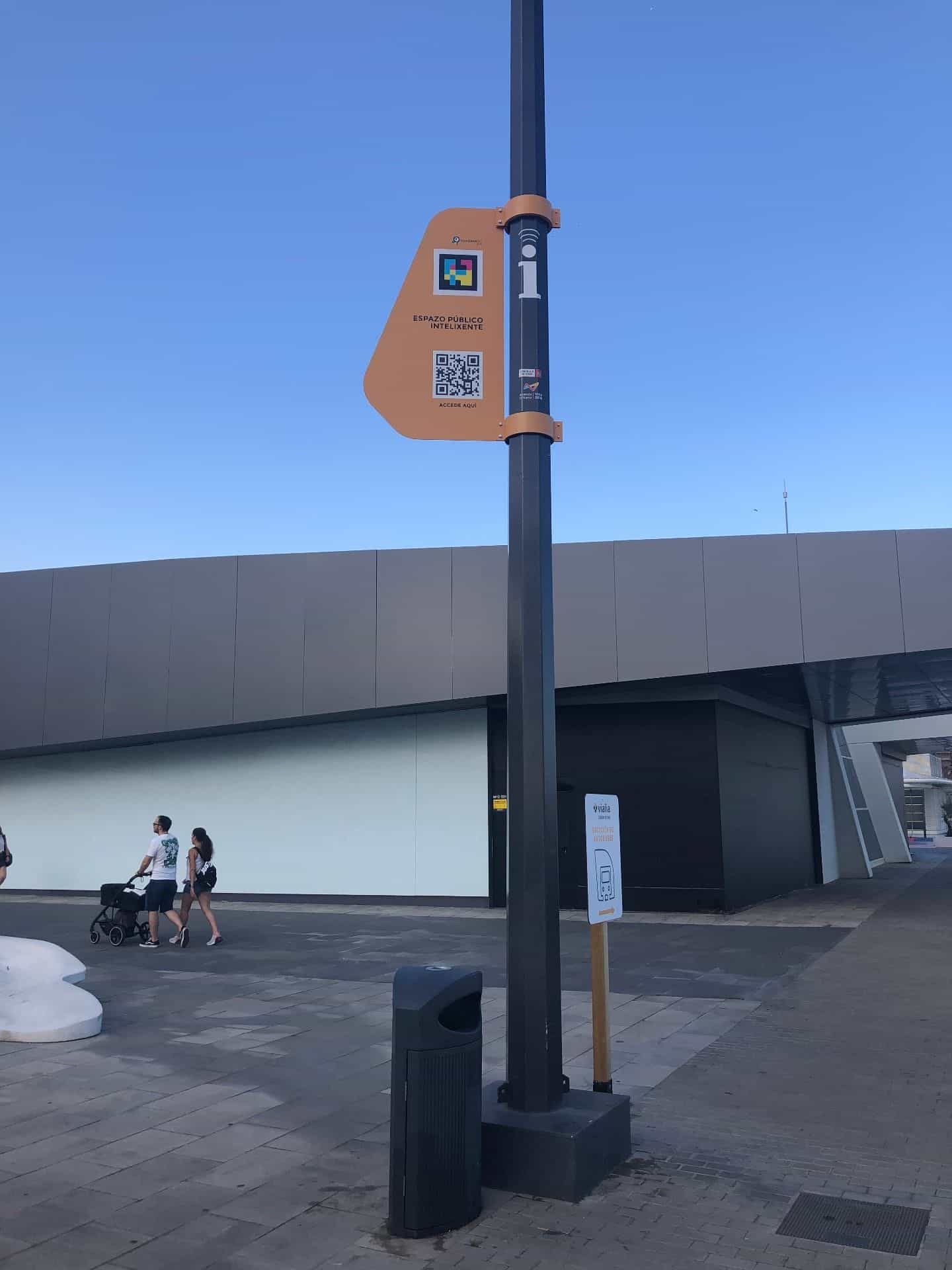 Spiazzo esterno della stazione intermodale Vialia Estación de Vigo con un palo nero di smart city su cui è appesa una targa arancione con il logo NaviLens GO, il suo codice a colori e il QR di accesso all’Espazo Público Intelixente; in fondo, una famiglia cammina con il passeggino e un cartello segnala la «Estación de autobuses»
