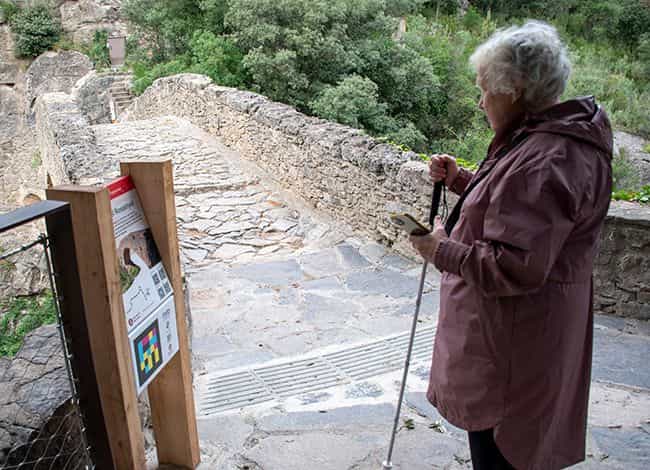 Eine Besucherin mit weißem Langstock und Handy in der Hand vor der Tafel „Rossinyol“ auf Holzpfosten mit ihrem NaviLens-Code, auf dem gepflasterten Weg der Rossinyol-Brücke in Sant Miquel del Fai; am Boden ein Streifen Bodenindikatoren