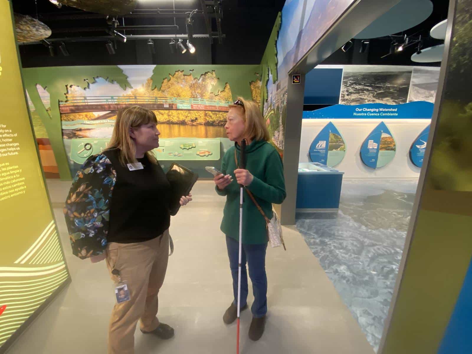 A visitor with a white cane talking to a WaterShed educator in front of the “Our Changing Watershed / Nuestra Cuenca Cambiante” panel, with its information droplets on urbanization and water quality