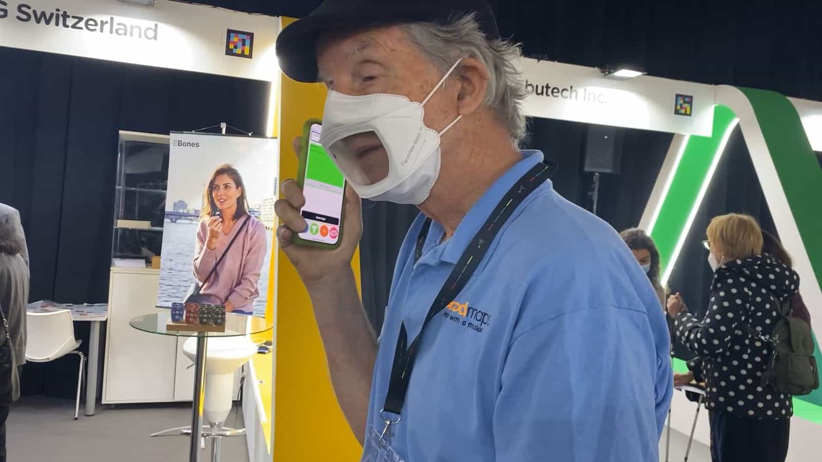 Visitatore anziano con polo azzurro della marca «Good Maps» e mascherina bianca FaceView prova l’app NaviLens con un iPhone accanto agli stand di Bones AG Switzerland e Ambutech Inc., le cui testate portano il codice NaviLens; in fondo, un rollup di Bones e, a destra, l’arco verde luminoso della scenografia del quartiere fieristico