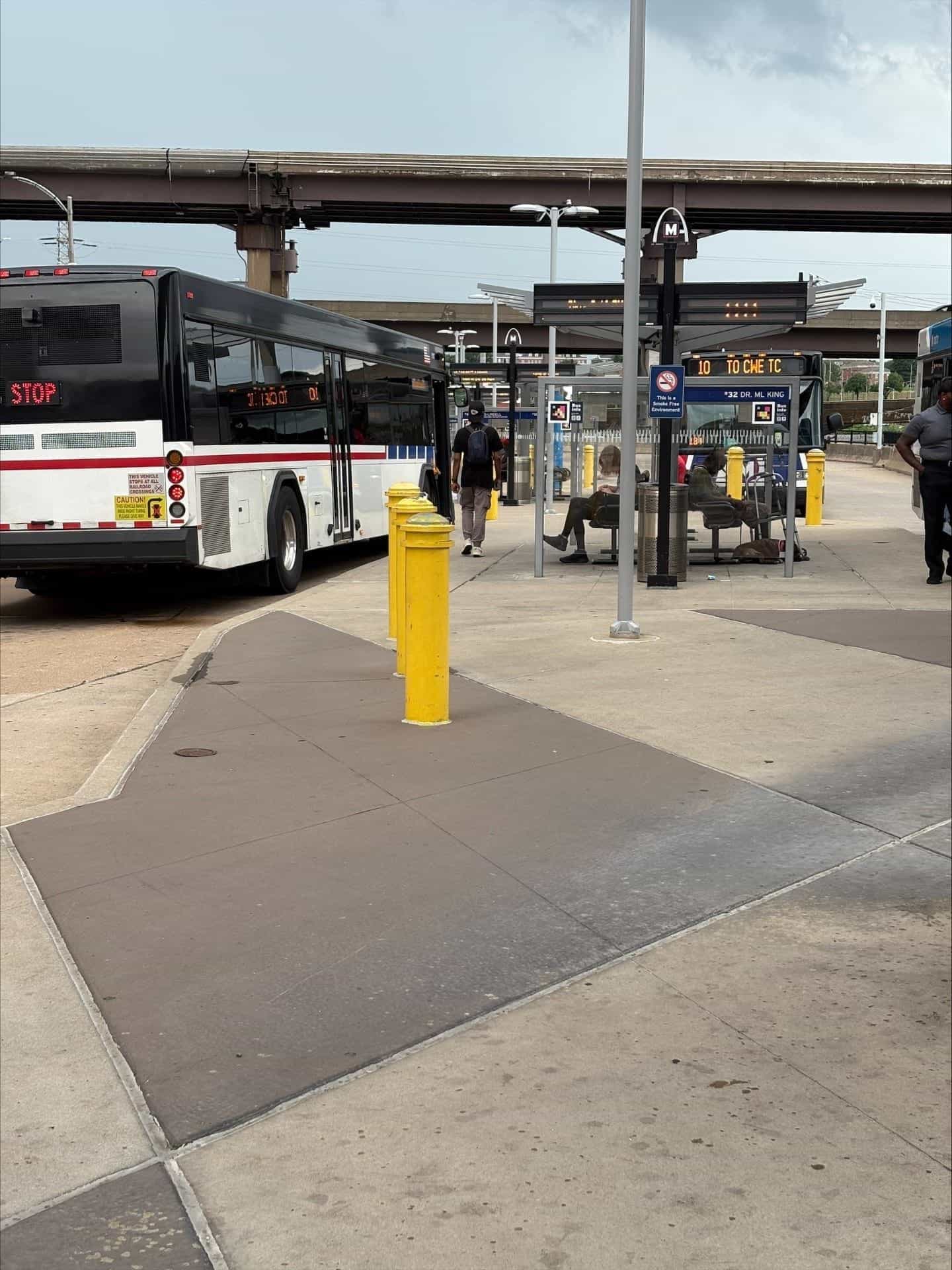 View of the Civic Center bus bays with the "10 TO CWE TC" bus on the left and the "32 DR. ML KING" bus on the right; in the middle, a person with a backpack, cap and grey trousers walks between the shelter where two riders wait and the blue "Bus Info" sign with a NaviLens code; yellow bollards and lamp posts; the concrete elevated MetroLink walkway above