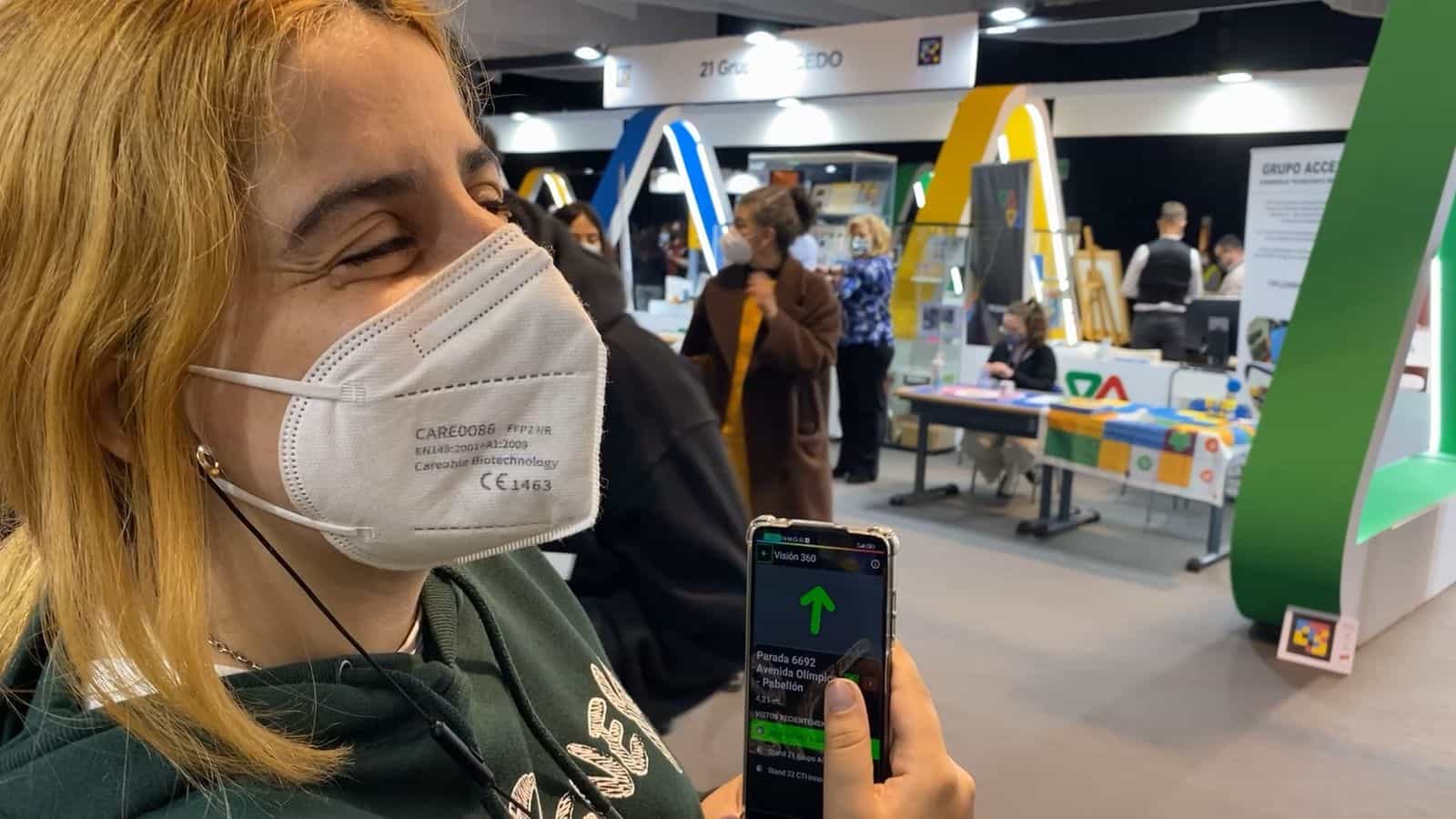 Giovane visitatore con felpa verde e mascherina bianca FFP2 tiene uno smartphone Android con l’app Vision 360º aperta: grande freccia verde che punta verso l’alto, testo «Fermata 6692 · Avenida Olímpica — Padiglione · 4,21 m» e, nell’elenco «Visite recenti», voci come «Stand 21 Grupo ACCEDO» e «Stand 22 CTI-Innov…»; in fondo, gli archi a forma di A azzurri, gialli e verdi del quartiere fieristico di Tifloinnova 2022