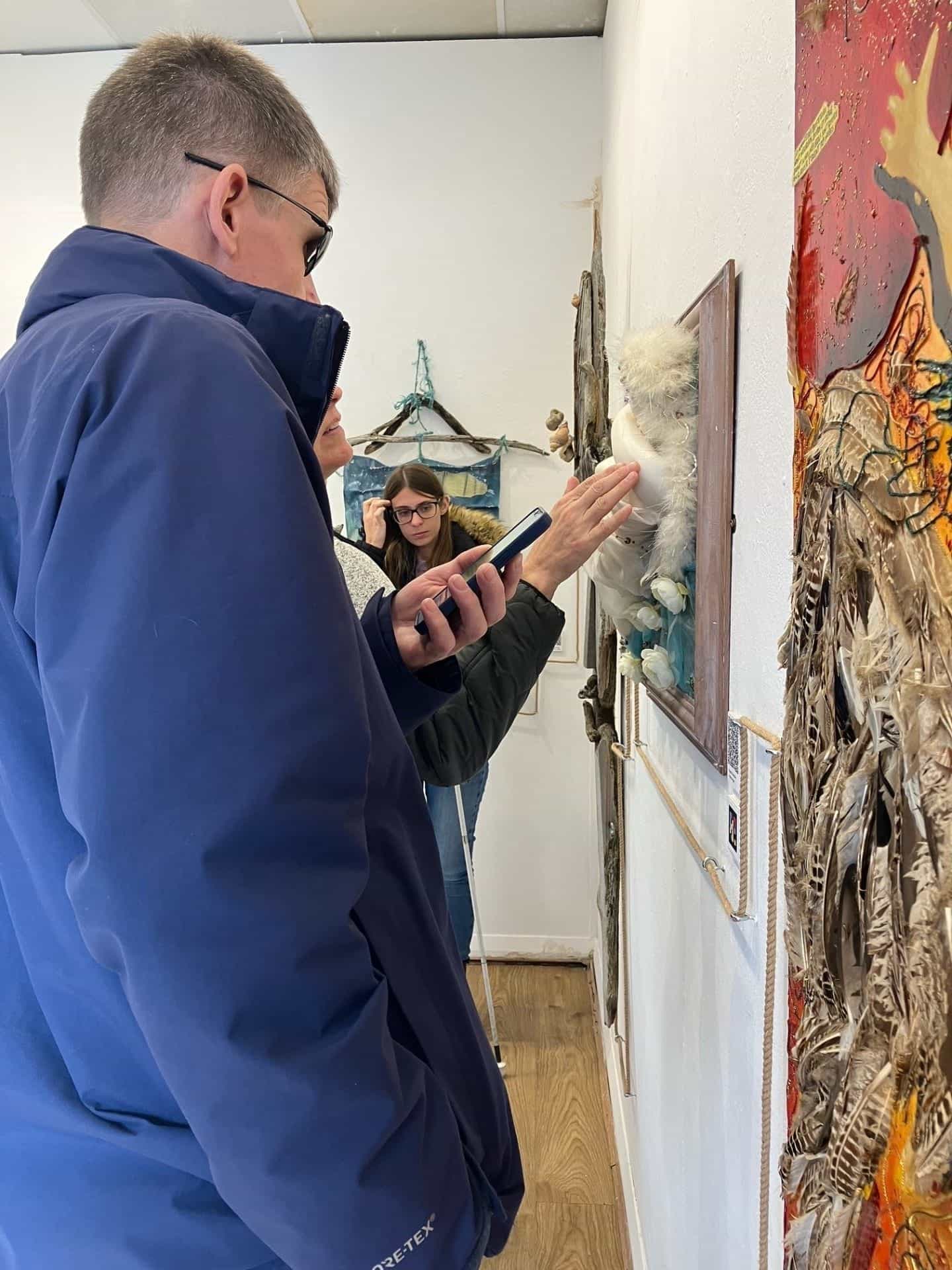 Due visitatori nella galleria davanti a un’opera di tecnica mista: in primo piano un uomo adulto con capelli corti bianchi, occhiali scuri e giacca Gore-Tex azzurra alza l’iPhone per inquadrare un codice NaviLens applicato alla parete; accanto a lui, una donna tocca con la mano destra una figura bianca con piume di un quadro tridimensionale incorniciato in legno; in fondo, un’altra visitatrice giovane con occhiali e giacca verde militare osserva la scena; di fianco si vede la corda guida tesa orizzontalmente sulla parete bianca
