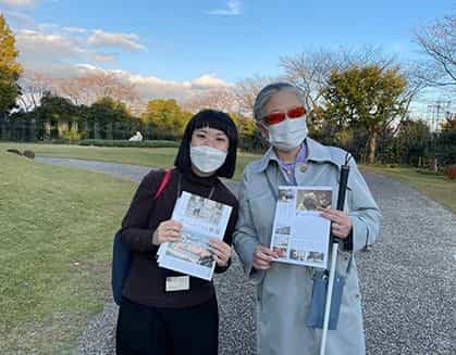 Zwei Besucherinnen des Museums — eine junge Kuratorin und eine ältere Frau mit Blindenstock und oranger Sonnenbrille — halten Flyer des Vangi-Museums im Außengarten in der Abenddämmerung