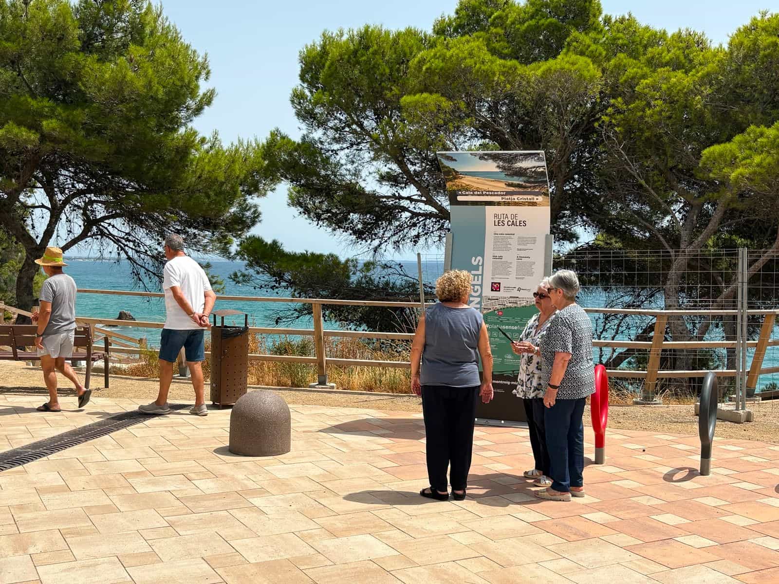 Drei Besucherinnen lesen mit dem Handy in der Hand das Totem der Cala dels Àngels auf einem gepflasterten Platz am Passeig de les Cales, dahinter Pinien und Meer