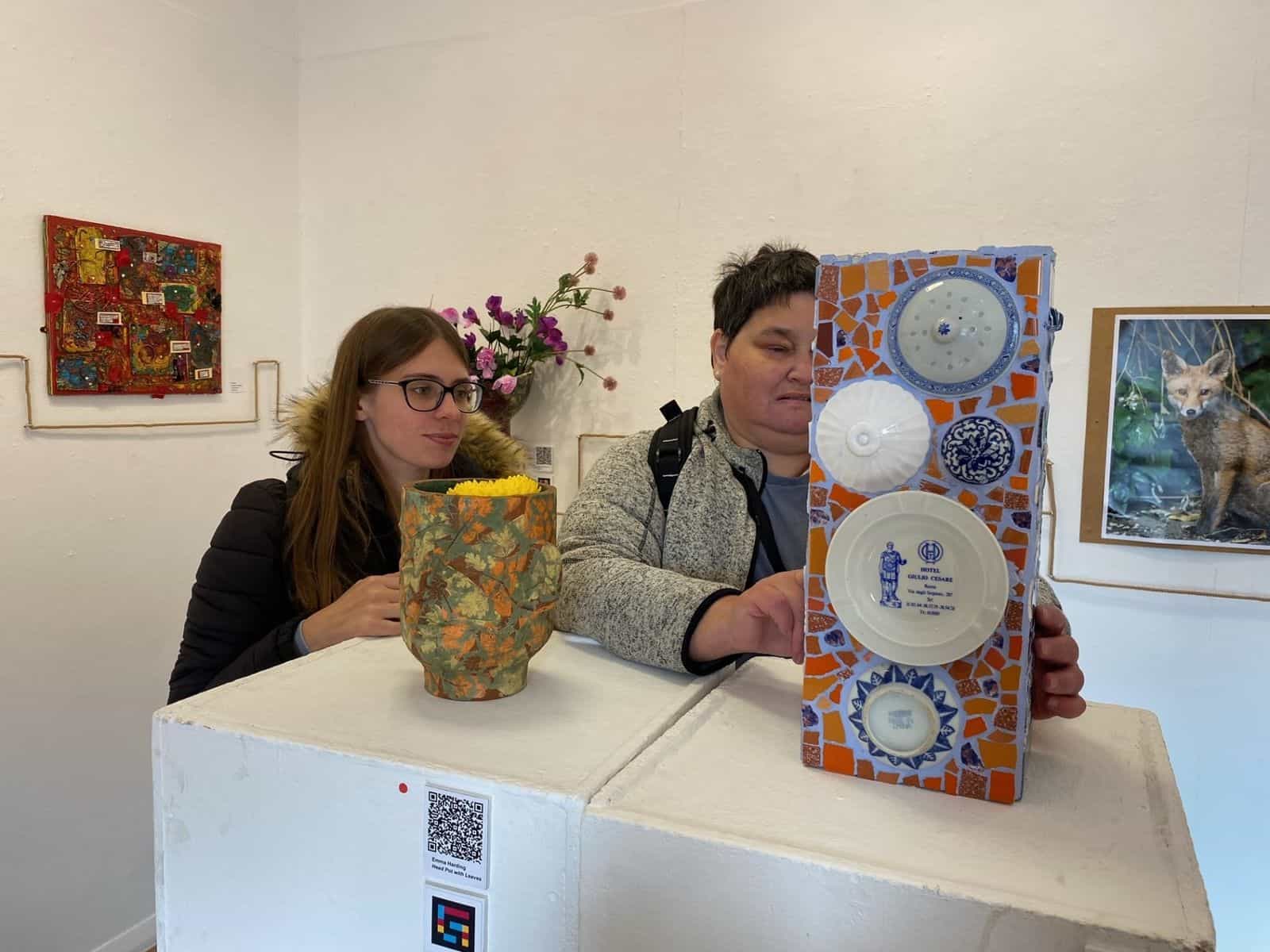 Due visitatrici sedute dietro un basamento bianco dentro la galleria con un vaso di ceramica decorato con foglie di quercia e un mosaico verticale di frammenti di piatti azzurri e bianchi su fondo arancione; sul basamento, una didascalia «Emma Harding — Head Pot with Leaves» con codice NaviLens