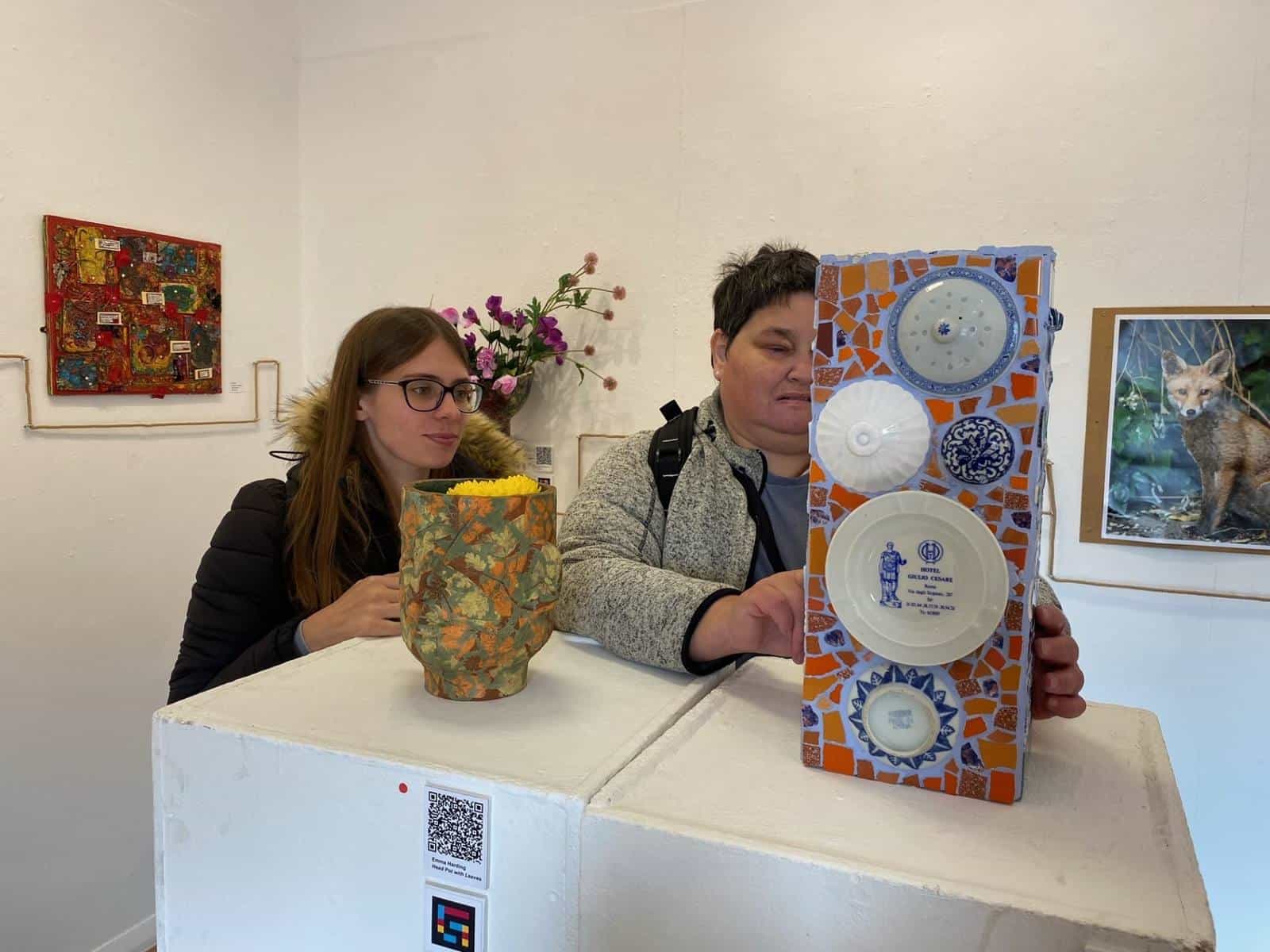 Two visitors seated behind a white plinth inside the gallery with a ceramic vessel decorated with oak leaves and a vertical mosaic of blue and white plate fragments on an orange background; on the plinth, a label “Emma Harding — Head Pot with Leaves” with a NaviLens code