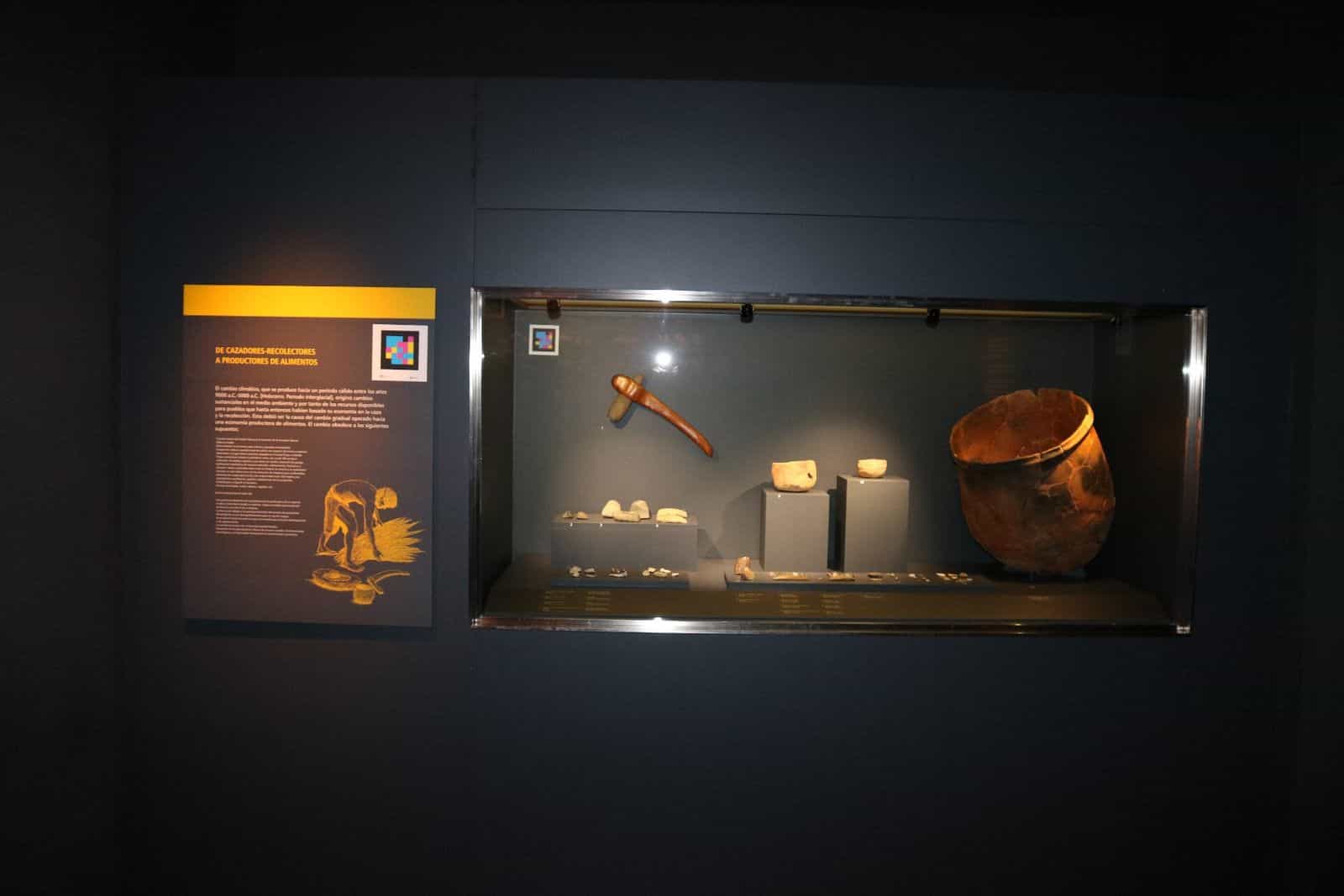 Vitrine „Von Jägern und Sammlern zu Nahrungsmittelproduzenten“ im MaYe Yecla, mit prähistorischem Gefäß, Holzsichel, Handmühlen und NaviLens-Code auf der Tafel und in der Vitrine