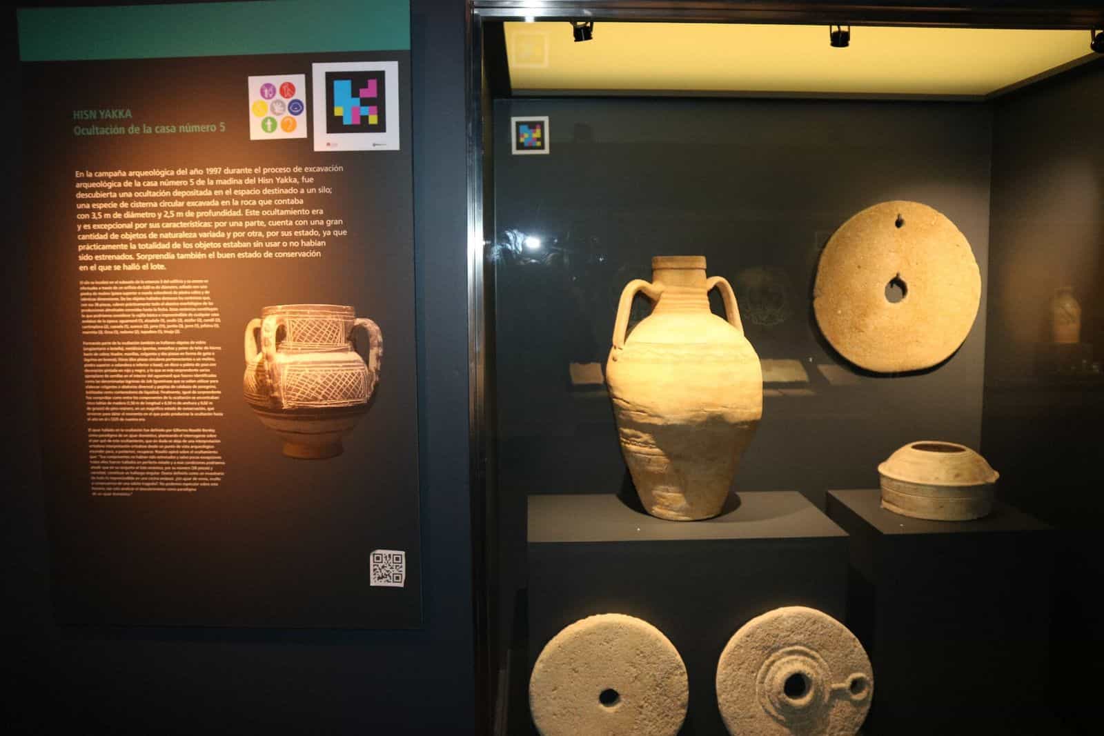 Vitrine „Hisn Yakka – Hortfund Haus Nr. 5“ im MaYe Yecla mit andalusischem Krug, Mühlen und verzierter Keramik, Tafel mit Barrierefreiheits-Piktogrammen und NaviLens-Code