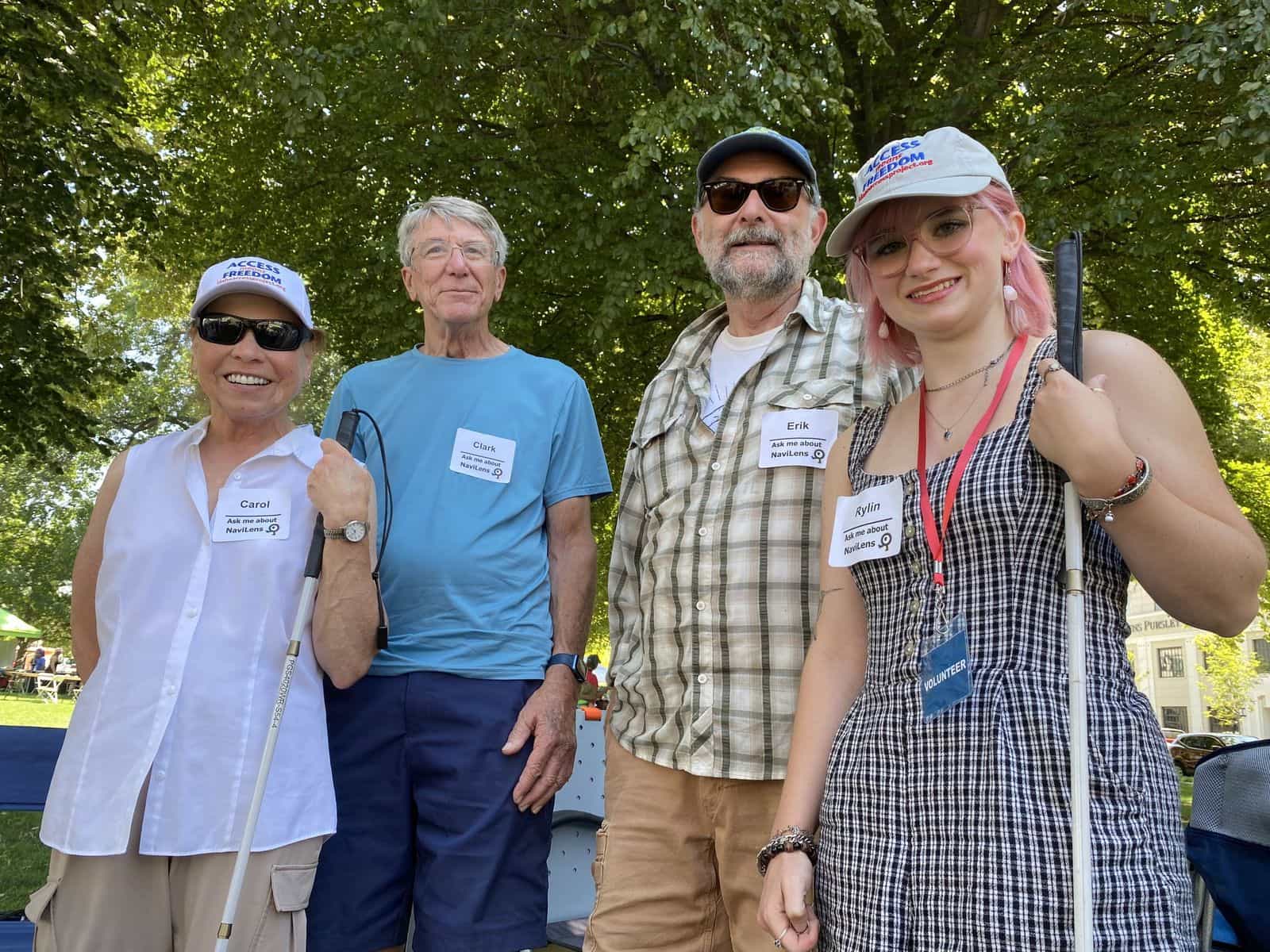 Vier Freiwillige von Hands Around the Capitol mit „Access = Freedom“-Kappen und Namensaufklebern mit dem Text „Ask me about NaviLens“; zwei von ihnen halten einen Blindenstock