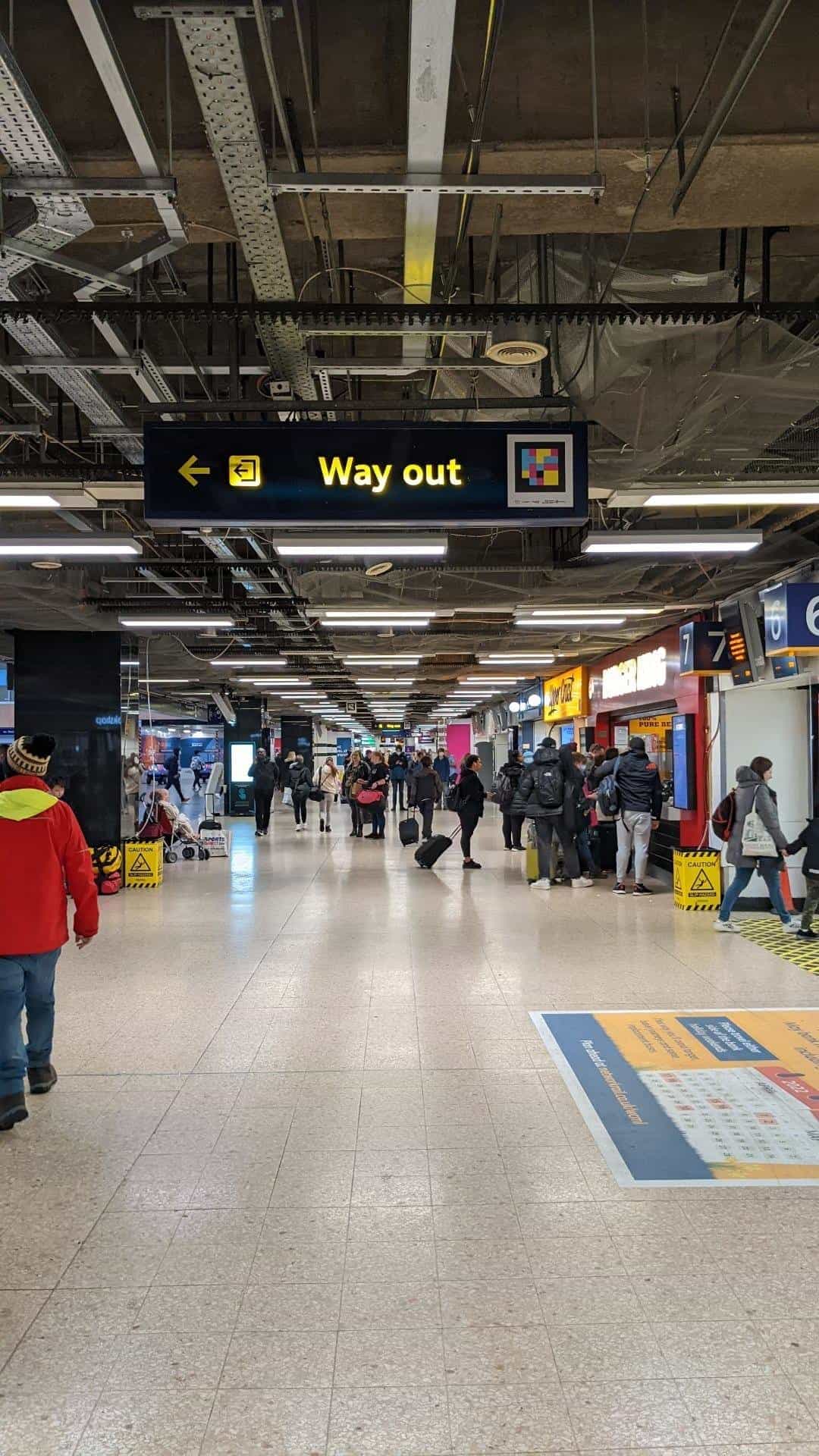 Suspended «Way out» sign with yellow exit arrow and integrated NaviLens code — Euston concourse with Burger King and platforms 6-7 in the background