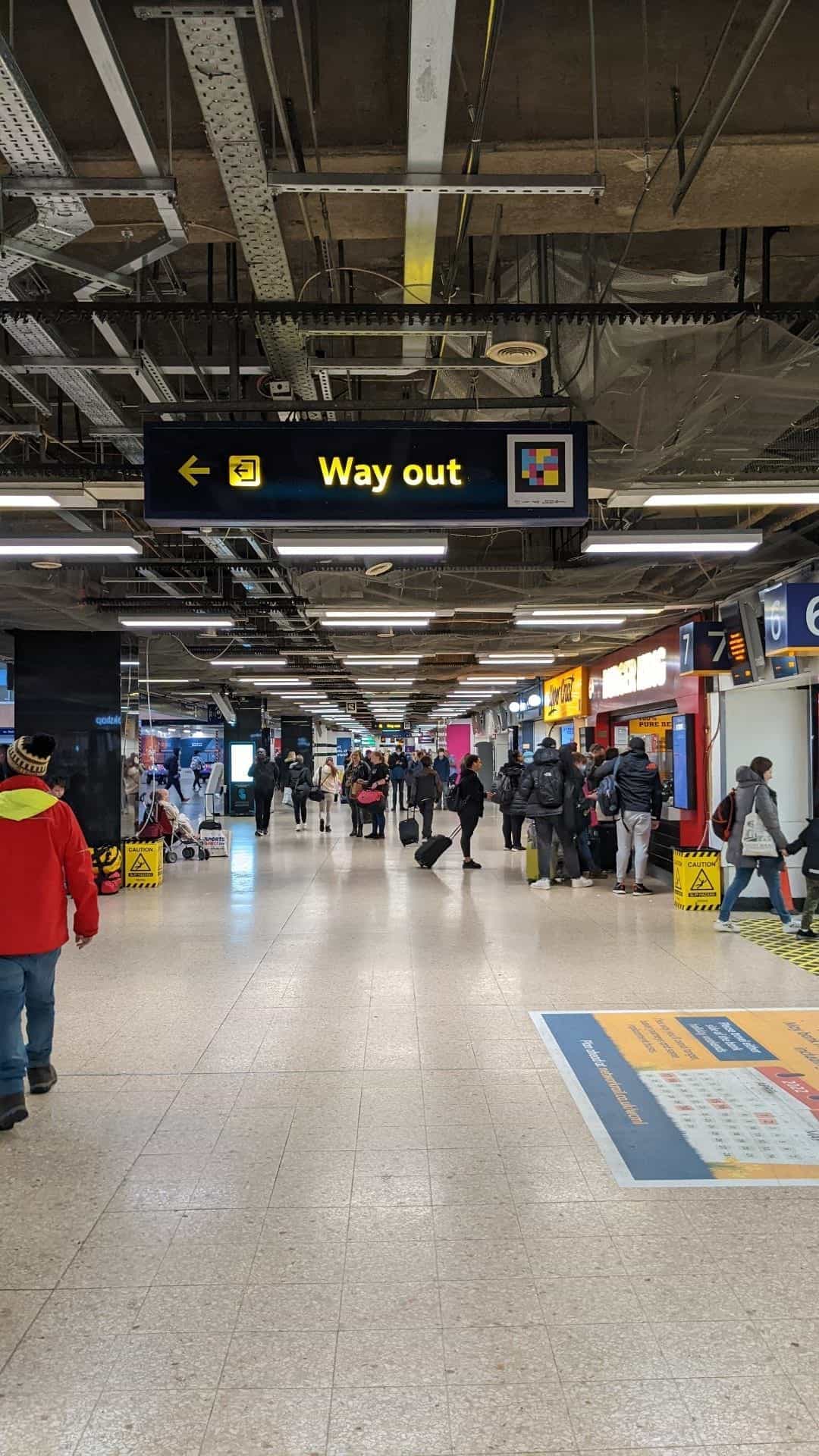 Suspended «Way out» sign with yellow exit arrow and integrated NaviLens code — Euston concourse with Burger King and platforms 6-7 in the background