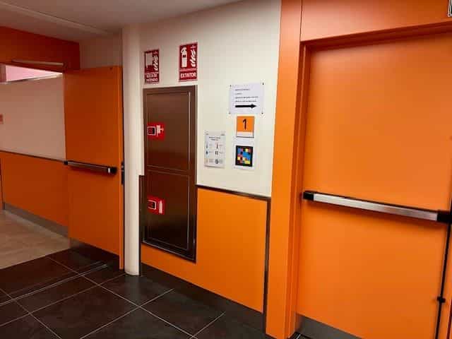 Zone 1 im CRE ONCE Madrid: weiße Wand mit nummeriertem Schild „1“, einer Karte mit barrierefreiem QR-Code und Richtungsangaben neben einer Feuerlöschersäule; im Hintergrund orangefarbene Wände und dunkler Steinzeugboden