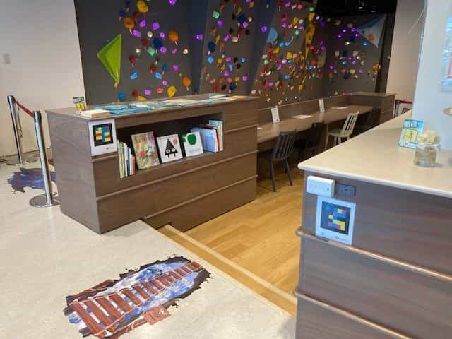 ビジョンパークのキッズエリア。絵本、木製テーブル、カラフルなホールドのクライミングウォール。本棚の正面とサイドキャビネットの側面に1枚ずつ、計2つのNaviLensタグが見える