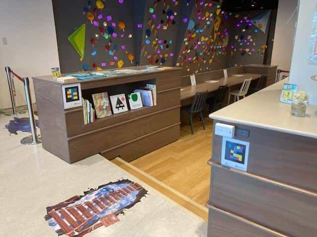 Children's area in the Vision Park with picture books, wooden table and a climbing wall with colourful holds; two NaviLens tags visible — one on the front of the bookcase and one on the side of the auxiliary unit